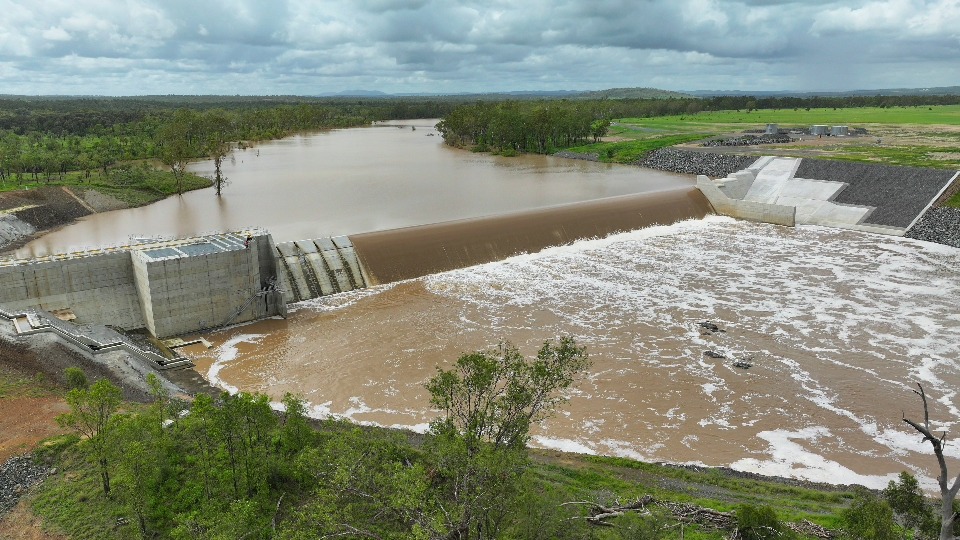 Significant volumes of water flowing from the Fitzroy River in the background, spilling over the completed Rookwood Weir and creating a whitewash effect as the water continues to flow down river from the weir. The river is surrounded by green pastural land.