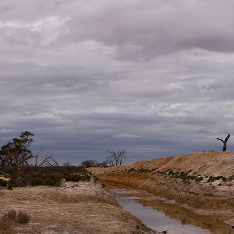 A muddy field with a floodwater river running through with storm clouds above.