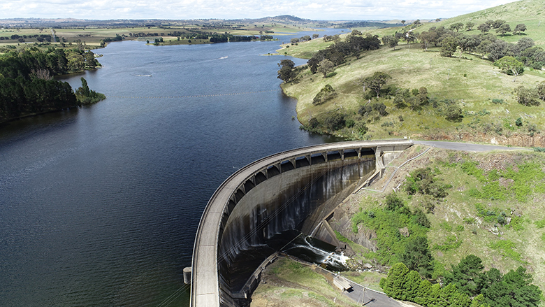 An aerial view of the Carcoar Dam on the Belubula River. The curved concrete dam wall is in the foreground, with green hills scattered with trees on the right side of the dam.