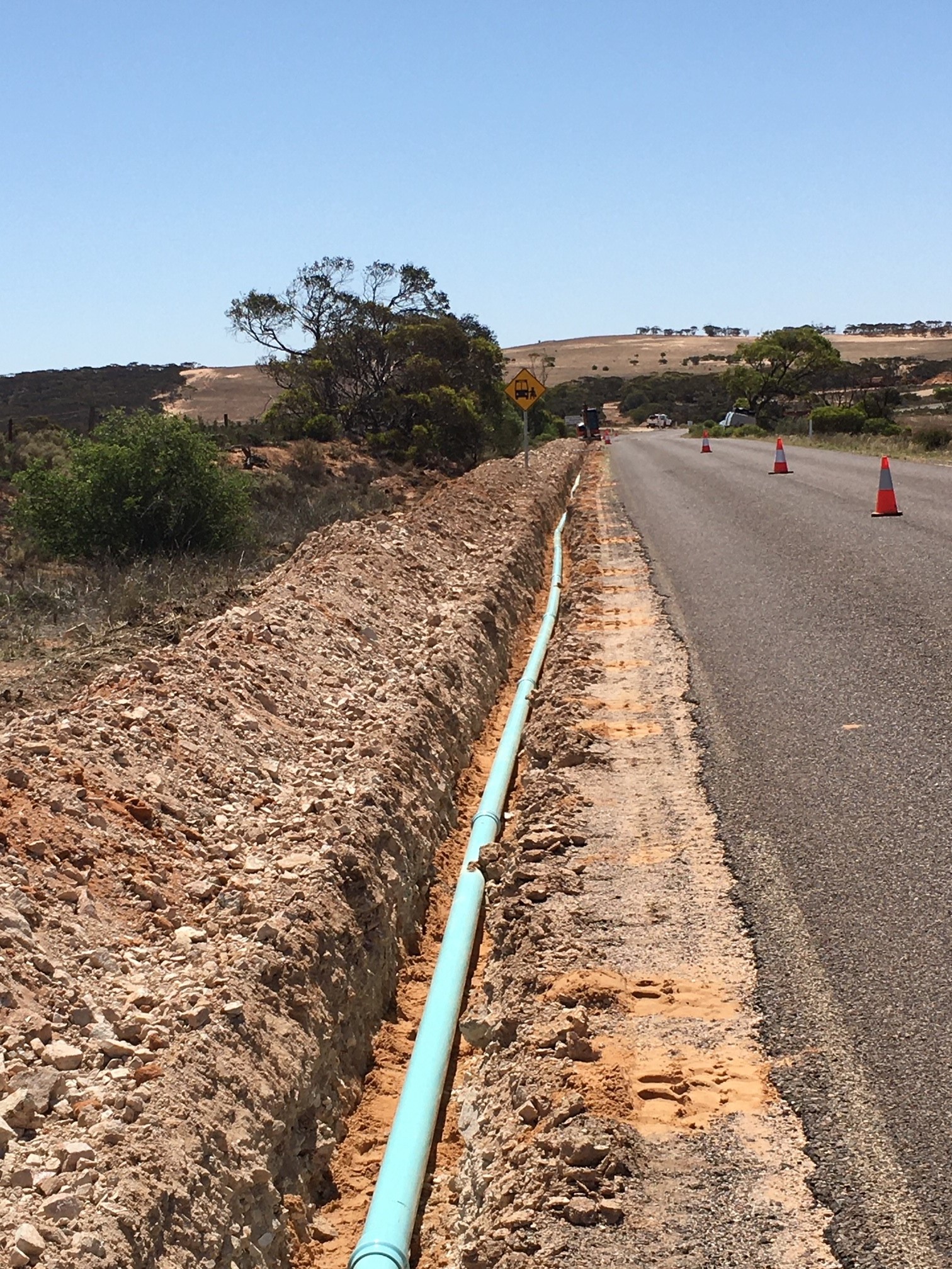 Laying pipe at Coolanie Water Scheme in South Australia. The light blue pipe is laid in a trench in the centre of the image, to the left of a country road.