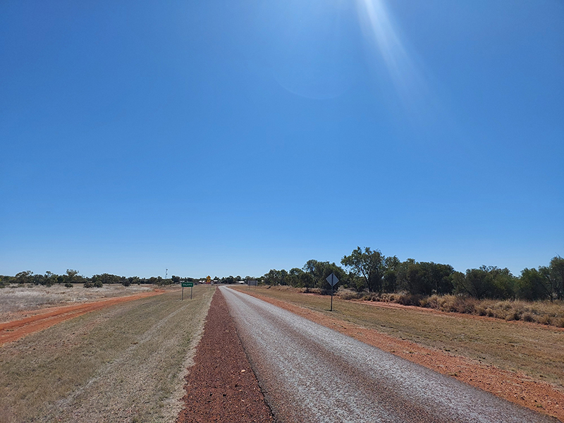 Single-lane road in the outback leading into the Dajarra community, with a blue sky. The road is surrounded by grass, red dirt and scrubby vegetation.