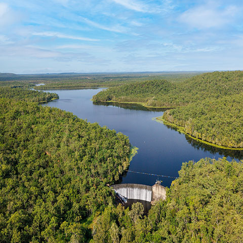 Image of Manton Dam, surrounded by green bush land, with a semi-circular concrete dam wall in the foreground and blue sky and clouds above. 