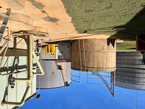 Several concrete water tanks with ladders and piping. There is grass and concrete in the foreground and blue sky above the tanks. 