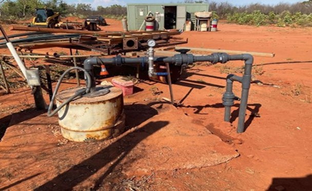 A bore sitting on an aged concrete pad with grey pipes running between the top of the bore and the ground beside it. A green shed and tractor sits in the background with scrubby bush behind them. Everything is sitting on and surrounded by red dirt.