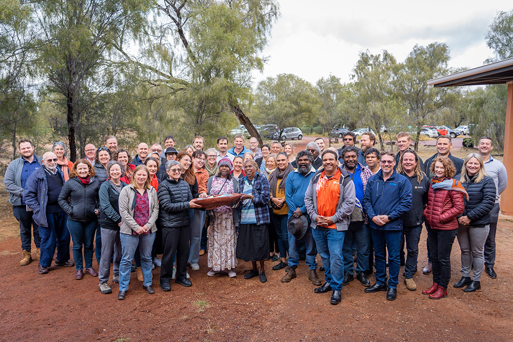 A group of people standing outside with the 3 people in the front holding a coolamon (wooden carrying vessel)