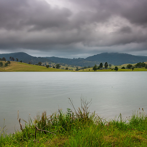 Lostock Dam with green grass in the foreground and green hills in the background with a cloudy sky above.