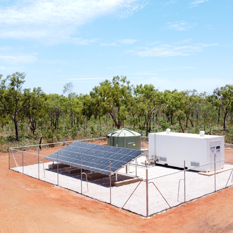 An example of a mini plant compound. A concrete pad surrounded by fencing with a water storage tank, solar panels and shipping container that houses water treatment equipment. There is red dirt surrounding the compound and native trees behind the compound. 