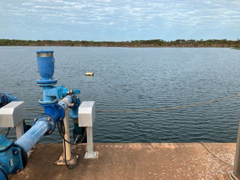 Dithery Creek Dam with dark blue water and bright blue dam equipment, including a water pump and pipes. There is a concrete dock in the foreground where the equipment sits. A blue cloudy sky and green vegetation is in the background on the horizon.