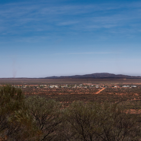 Looking across a desert landscape in the Northern Territory, with native trees and bushes in the foreground and a remote community in the distance. 