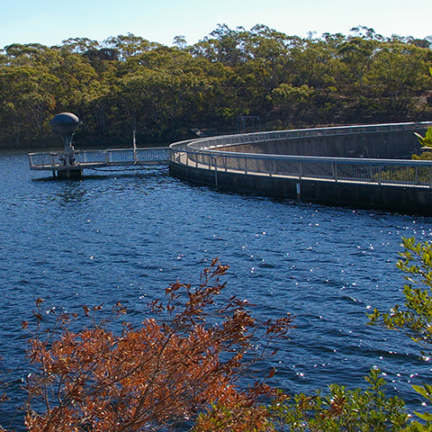 Large dam with short dam walls on the right and bushland in the background, sunny day.