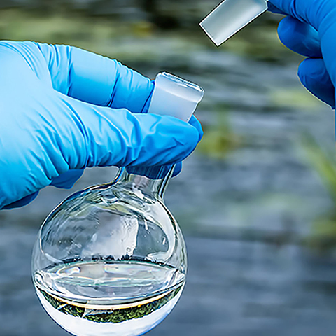 Close up of hands wearing blue gloves holding a small beaker with a water sample, and a river in the background.