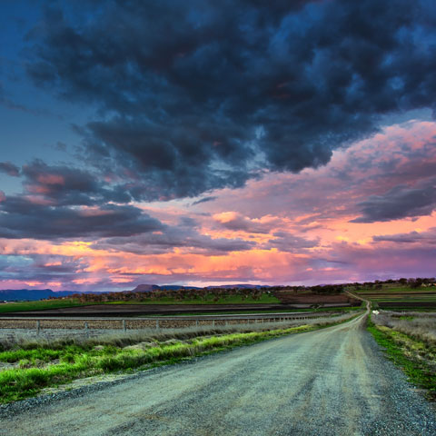 A gravel and dirt road through farmland below a cloudy pink sunset.