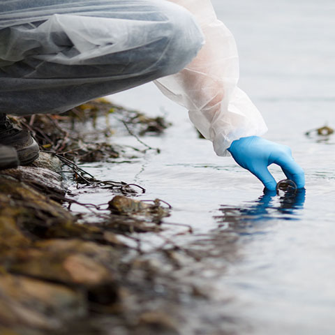 Person in protective clothing and blue gloves gathering a water sample from the rocky bank of a body of water.