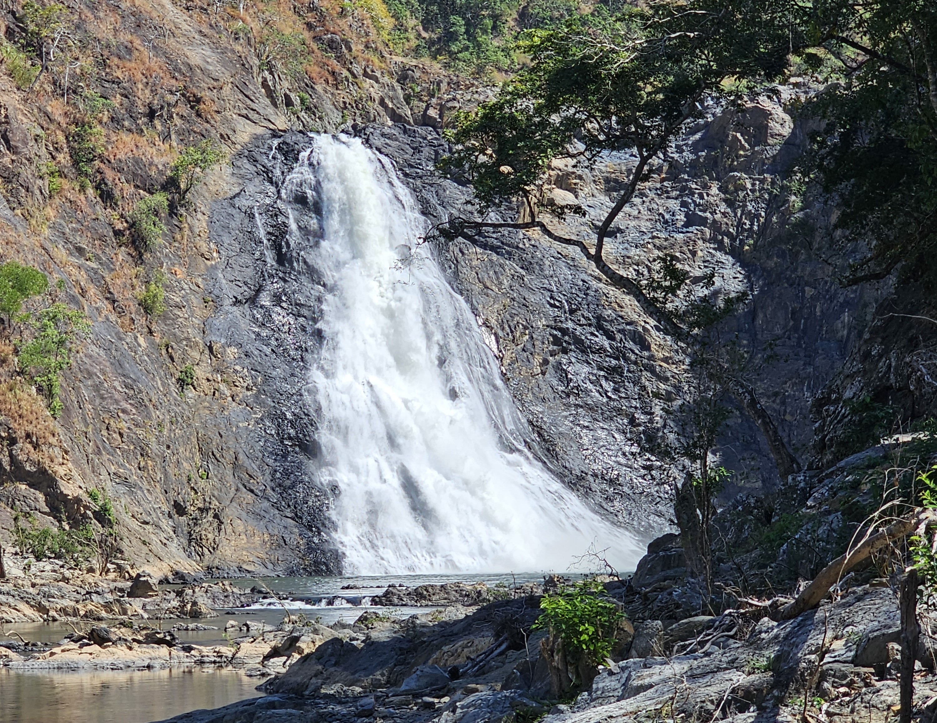 Waterfall surrounded by steep, dark, rocky cliffs and trees. There is a bright blue sky above the waterfall. 